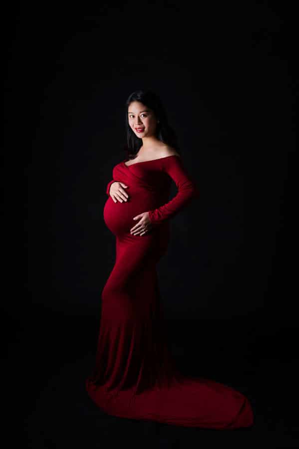 full length maternity portrait of pregnant Asian woman in red gown in studio on dark backdrop
